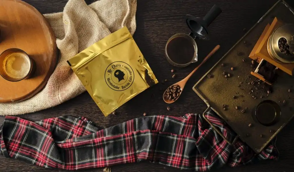 Bolsa dorada de café Mr. Chava sobre madera, con molinillo y granos tostados